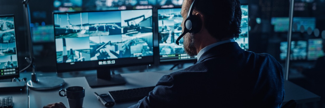 Male Officer Works on a Computer with Surveillance CCTV Video in a Harbour Monitoring Center with Multiple Cameras on a Big Digital Screen. Employees Sit in Front of Displays with Big Data.